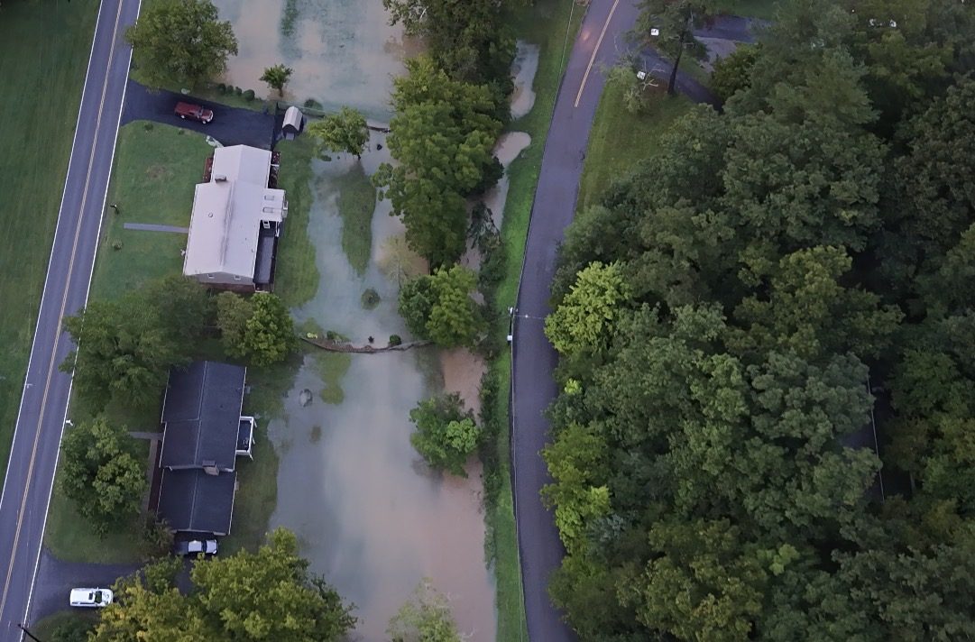 Top-down drone photo of floodwater covering yards and a driveway beside a rural highway after heavy rain